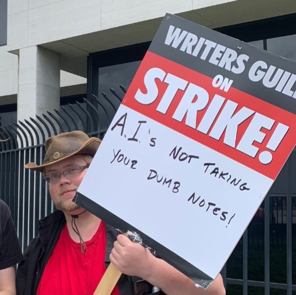 A picketer, David James Henry, carries a sign that reads "A.I.'s not taking your dumb notes!"